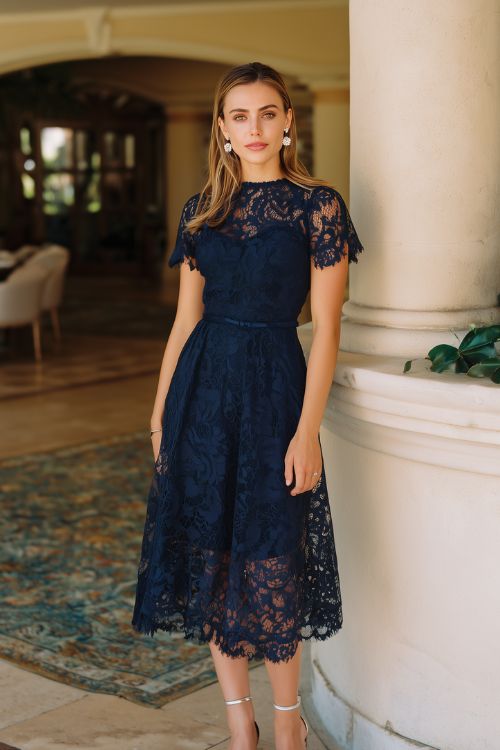 A woman wears a navy blue lace wedding guest dress with short sleeves, a high neckline, and a tea-length hem