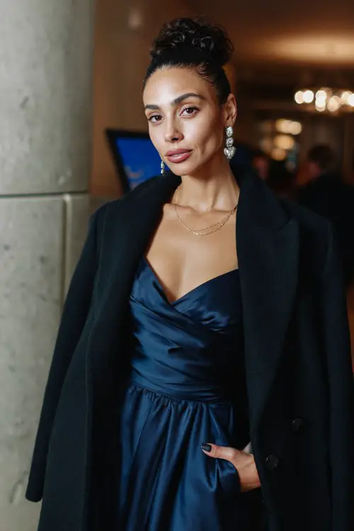 A woman wears a navy satin wrap gown, paired with a black wool coat and silver drop earrings, at an elegant city venue