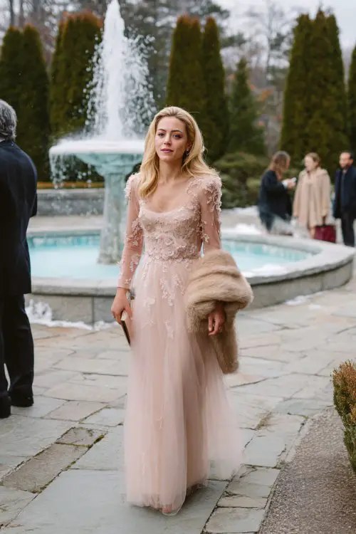 A woman wears a pastel pink long-sleeve tulle gown with embroidered floral appliqués, layered with a soft faux fur capelet