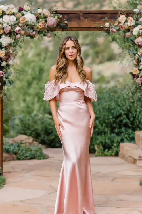A woman wears a pink floor-length satin mermaid gown with an off-the-shoulder neckline and pearl-embellished cape sleeves