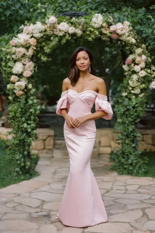 A woman wears a pink floor-length satin mermaid gown with an off-the-shoulder neckline and pearl-embellished puff sleeves