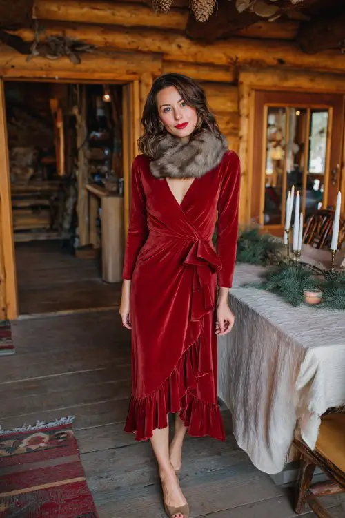 A woman wears a red long-sleeve wrap dress with velvet texture and ruffled hem, paired with a faux fur scarf and neutral heels