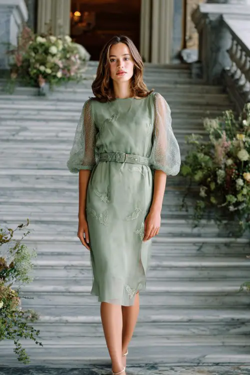 A woman wears a sage green slip dress with sheer embroidered sleeves and a structured belt, stepping into a marble hall