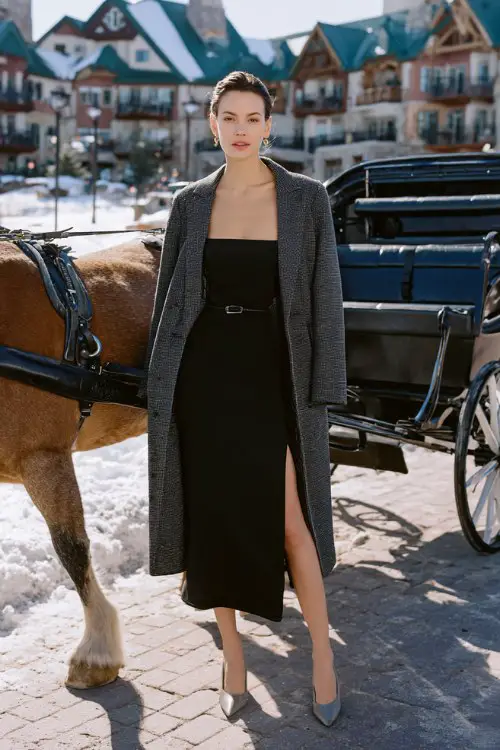 A woman wears a sleek black column dress with a square neckline and side slit, styled with a belted charcoal gray cashmere coat and pointed-toe heels