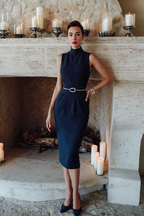 woman over 40 wears a fitted navy midi dress with a high neckline and a pearl belt, paired with pumps
