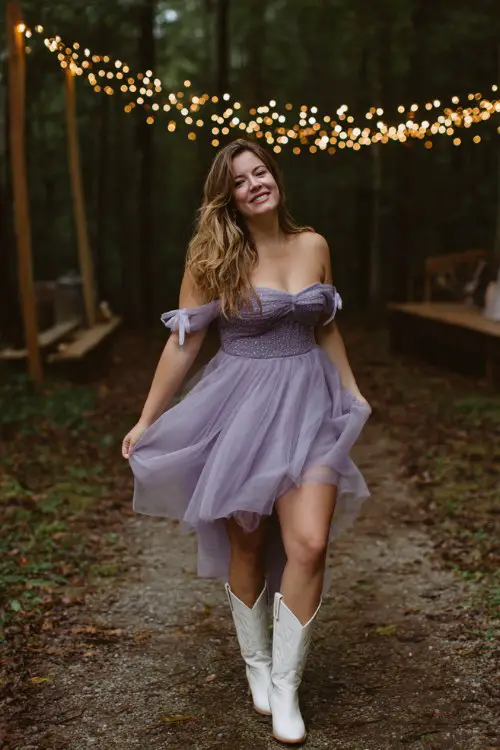 A curvy woman wears a lilac layered tulle midi dress with off-the-shoulder bows and sequin detailing, paired with white cowgirl boots