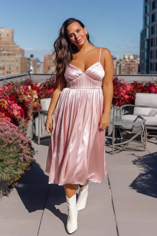 A curvy woman wears a pink satin midi dress with a pleated skirt and sweetheart neckline, paired with white ankle boots
