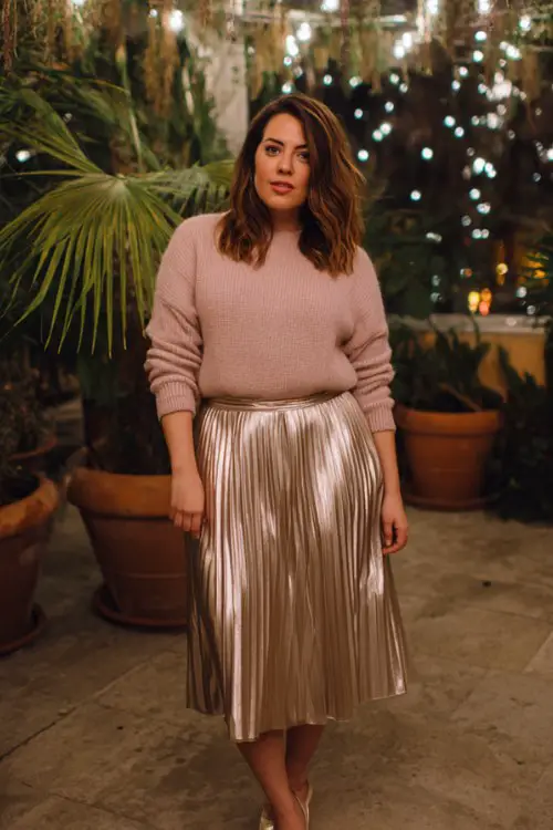 A curvy woman wears a soft blush sweater with a metallic pleated midi skirt and pointed heels