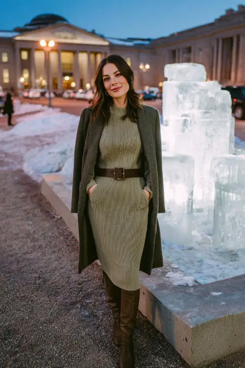 A curvy woman wears an olive green knit midi dress with a wide belt and knee-high boots, layered with a tailored wool coat