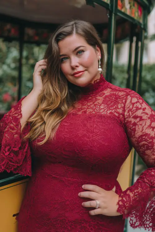 A full body of plus-size woman wears a bold cherry red high-neck lace dress with flared sleeves and pearl drop earrings