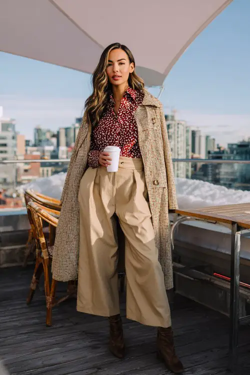 A petite woman wears a heart-print blouse layered under a structured cape coat, paired with wide-leg trousers and heeled boots