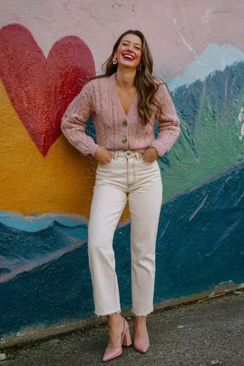 A petite woman wears straight-leg jeans, a pink cable-knit cardigan tucked in, heart-shaped earrings, and block heels