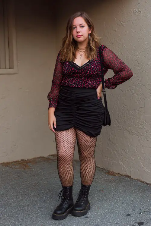 A plus-size woman layers a sheer cherry-print mesh top over a black bralette, tucked into a ruched mini skirt with fishnet tights and combat boots