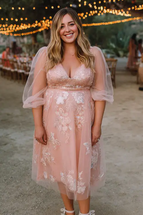 A plus-size woman wears a blush pink tulle midi dress with embroidered florals and sheer puff sleeves, paired with silver ankle-strap heels