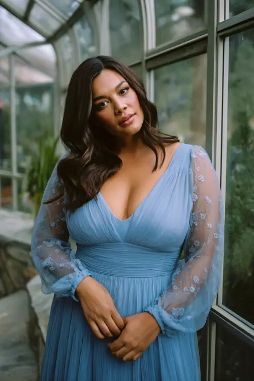 A plus-size woman wears a cornflower blue gown with an empire waist, pleated skirt, and sheer bishop sleeves embroidered with tiny florals