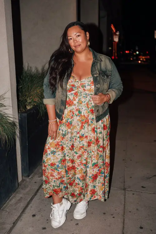 A plus-size woman wears a floral print maxi dress with a denim jacket and white sneakers for a movie night