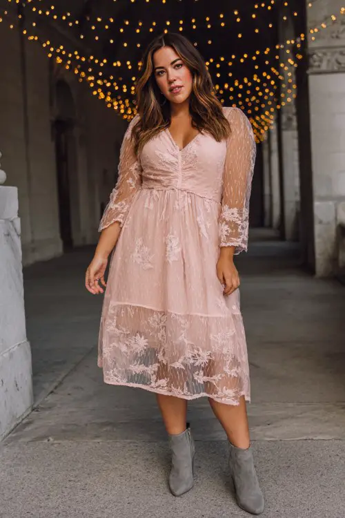 A plus-size woman wears a pastel pink midi dress with a sheer lace overlay and embroidered floral details, paired with soft gray ankle boots