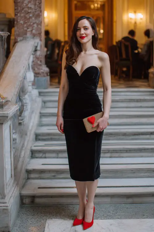 A woman over 30 wears a black velvet midi dress with a sweetheart neckline, styled with red heels and a clutch
