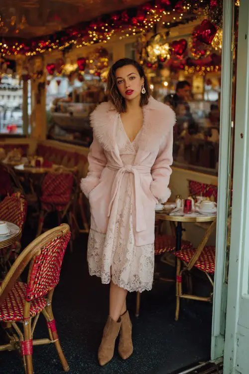 A woman over 30 wears a blush pink wrap coat with faux fur trim over a lace midi dress, suede ankle boots, and pearl earrings
