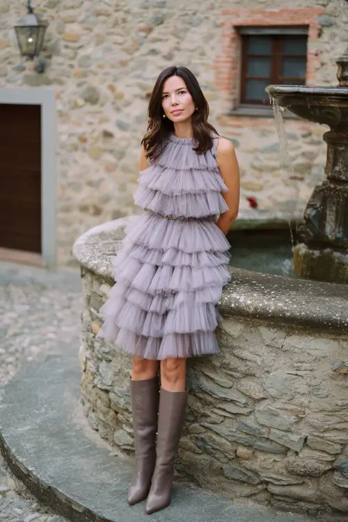 A woman over 30 wears a pale lavender tiered tulle dress with a ruffled neckline and taupe leather knee-high boots