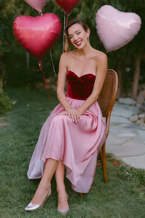 A woman over 30 wears a two-tone dress with a fitted velvet ruby-red bodice and a flowing pink chiffon skirt, styled with metallic heels