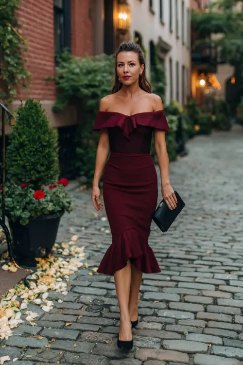 A woman over 40 wears a burgundy off-the-shoulder knee-length dress with a ruffled hem, styled with a black clutch and low heels