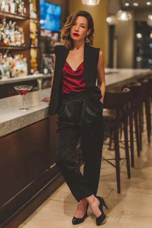 A woman over 40 wears a tailored black pantsuit with a silk red camisole underneath, red lipstick, and stiletto heels