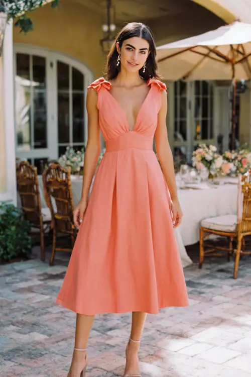 A woman wears a coral A-line midi dress with a plunging V-neckline, structured pleats, and bow-tie shoulders, styled with beige strappy sandals