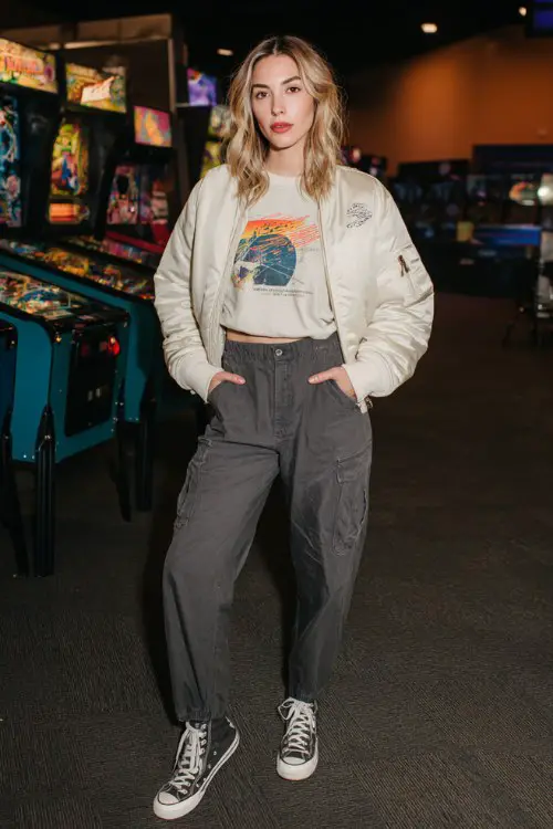 A woman wears a cream cropped bomber over a graphic tee, paired with charcoal cargo pants and high-top Converse