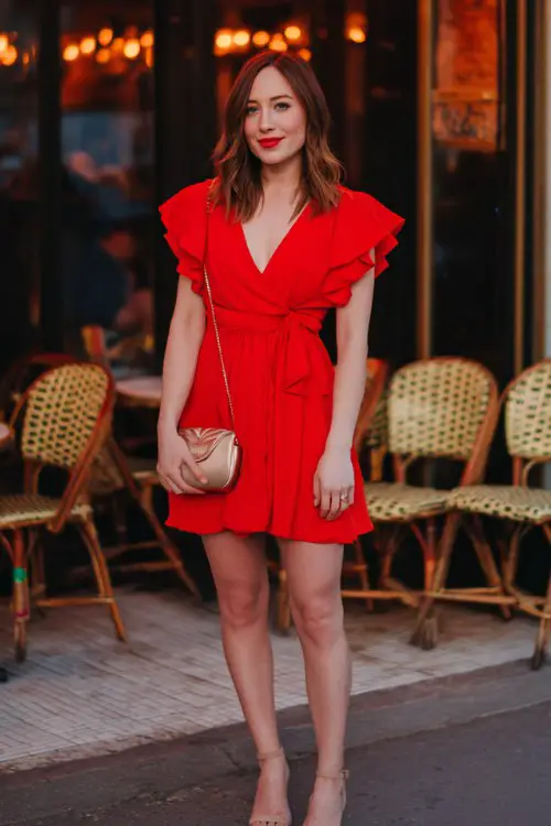 A woman wears a flirty red wrap dress with flutter sleeves, heels, and a heart-shaped clutch
