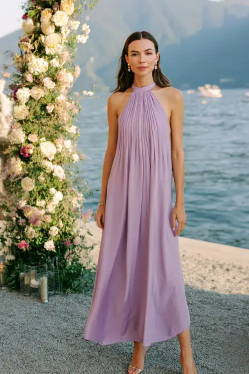 A woman wears a lilac pleated high-neck maxi dress with a keyhole back, styled with silver heels and pearl earrings