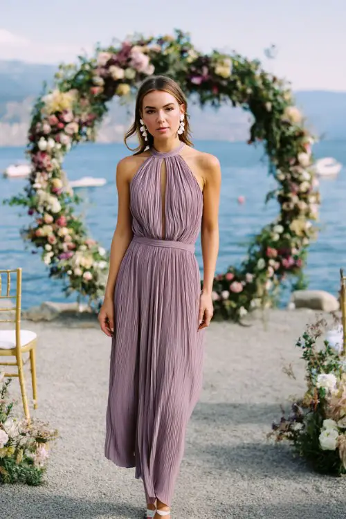 A woman wears a lilac pleated high-neck maxi dress with a keyhole back, styled with silver heels and pearl earrings