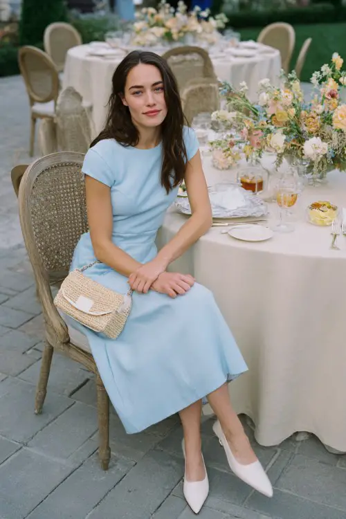 A woman wears a pale blue A-line midi dress with contrast stitching and scalloped cap sleeves, paired with soft white leather flats and a woven crossbody bag