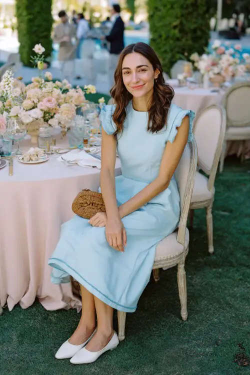 A woman wears a pale blue A-line midi dress with contrast stitching and scalloped cap sleeves, paired with soft white leather flats and a woven crossbody bag