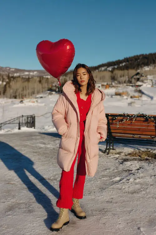 A woman wears a pale pink puffer coat over a red knit two-piece set and snow boots, holding a heart-shaped balloon
