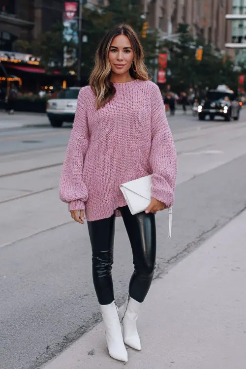 A woman wears a pink oversized knit sweater with faux black leather leggings and white ankle boots, and a white clutch