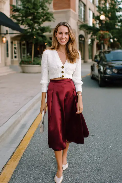 A woman wears a satin burgundy midi skirt, a cropped white cardigan with pearl buttons, and ballet flats