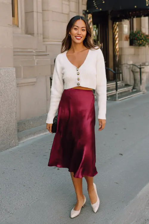 A woman wears a satin burgundy midi skirt, a cropped white cardigan with pearl buttons, and ballet flats