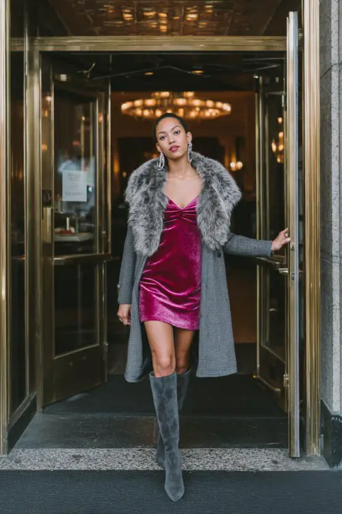 A woman wears a velvet deep pink mini dress under a dramatic longline gray coat with a fur collar, thigh-high boots, and bold earrings