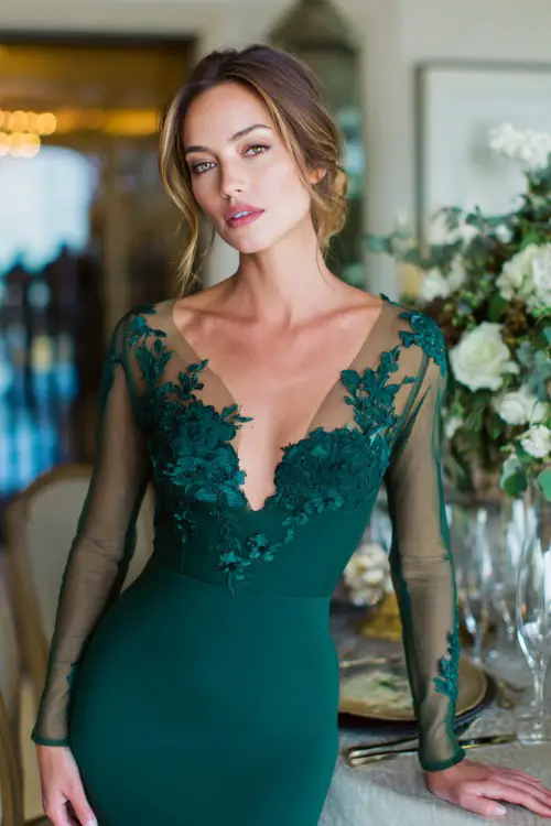 A woman wears an elegant deep emerald green gown with a fitted silhouette and sheer lace long sleeves, featuring a plunging illusion neckline with floral lace appliqué