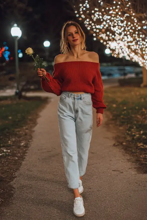 A woman wears light-wash straight-leg jeans, a soft red off-shoulder knit sweater, and white sneakers