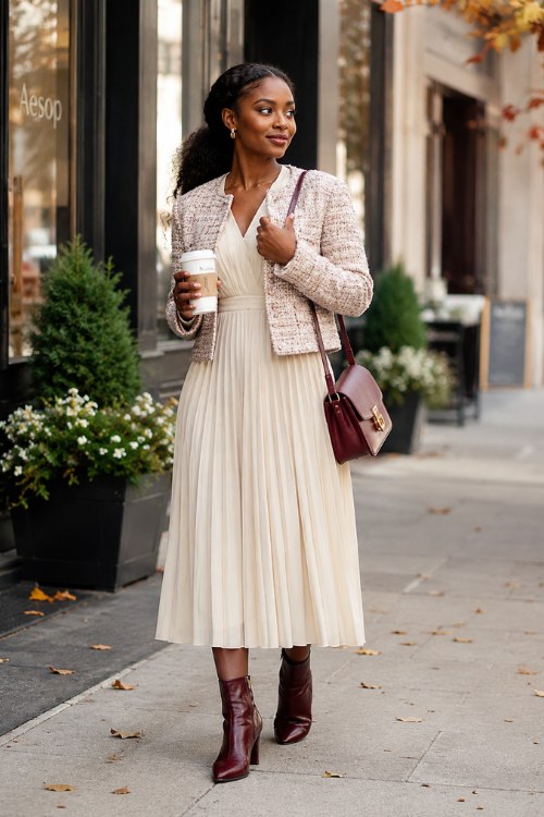 a Black woman wearing an ivory pleated midi dress layered under a cropped tweed jacket, paired with burgundy ankle boots, delicate gold earrings