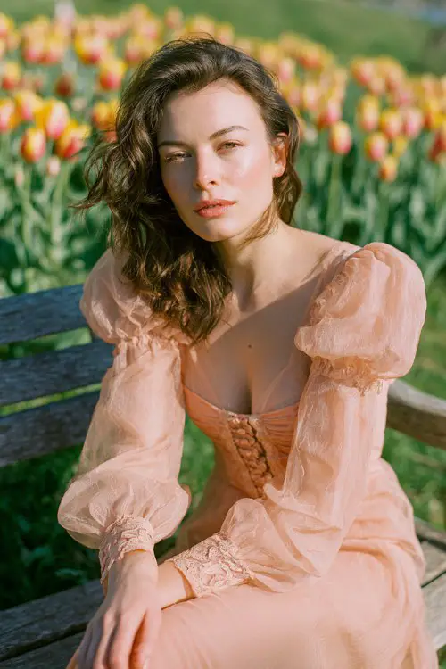 a woman sitting on a wooden bench in a field of blooming tulips, wearing a peach sheer vintage-style dress with lace details and puff sleeves