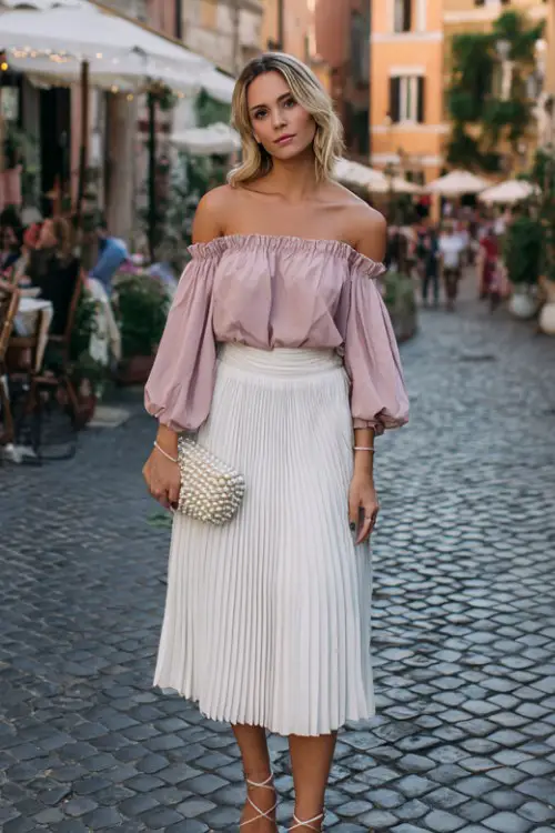 A woman over 30 wears a blush pink off-shoulder blouse tucked into a white pleated midi skirt, accessorized with lace-up sandals and a pearl-embellished clutch
