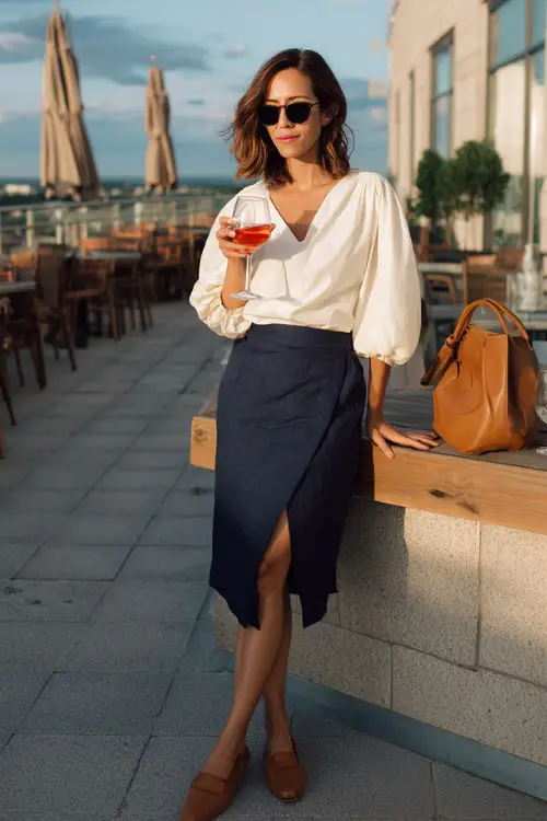 A woman over 40 wears a navy midi skirt with a side slit and a soft ivory blouse with gathered sleeves, paired with tan mules and a structured shoulder bag