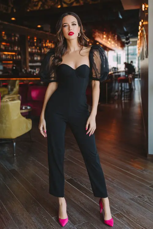 A woman wears a black fitted jumpsuit with sheer organza puff sleeves and a sweetheart neckline, styled with hot pink heels and bold red lips