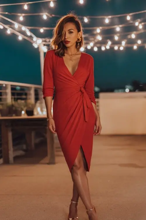 A woman wears a red wrap midi dress with heels and delicate gold jewelry, standing under a string of lights on a terrace