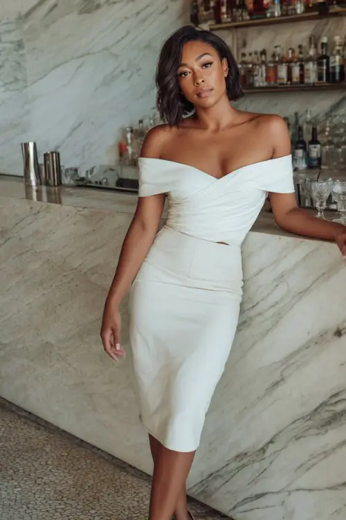 A Black woman wears a white off-shoulder top with a sleek pencil skirt and silver heels