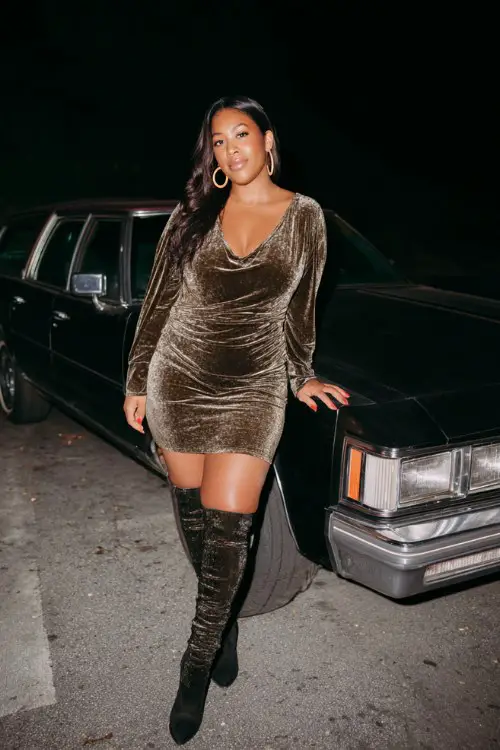 A curvy Black woman wears a velvet mini dress with long sleeves, gold hoops, and thigh-high boots