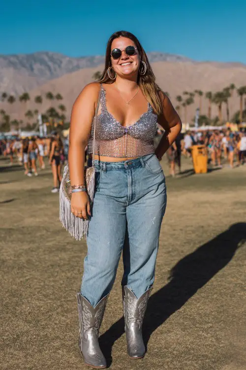A plus-size woman wears silver cowboy boots with high-waisted flare jeans and a rhinestone mesh crop top layered over a bralette, styled with bold hoop earrings